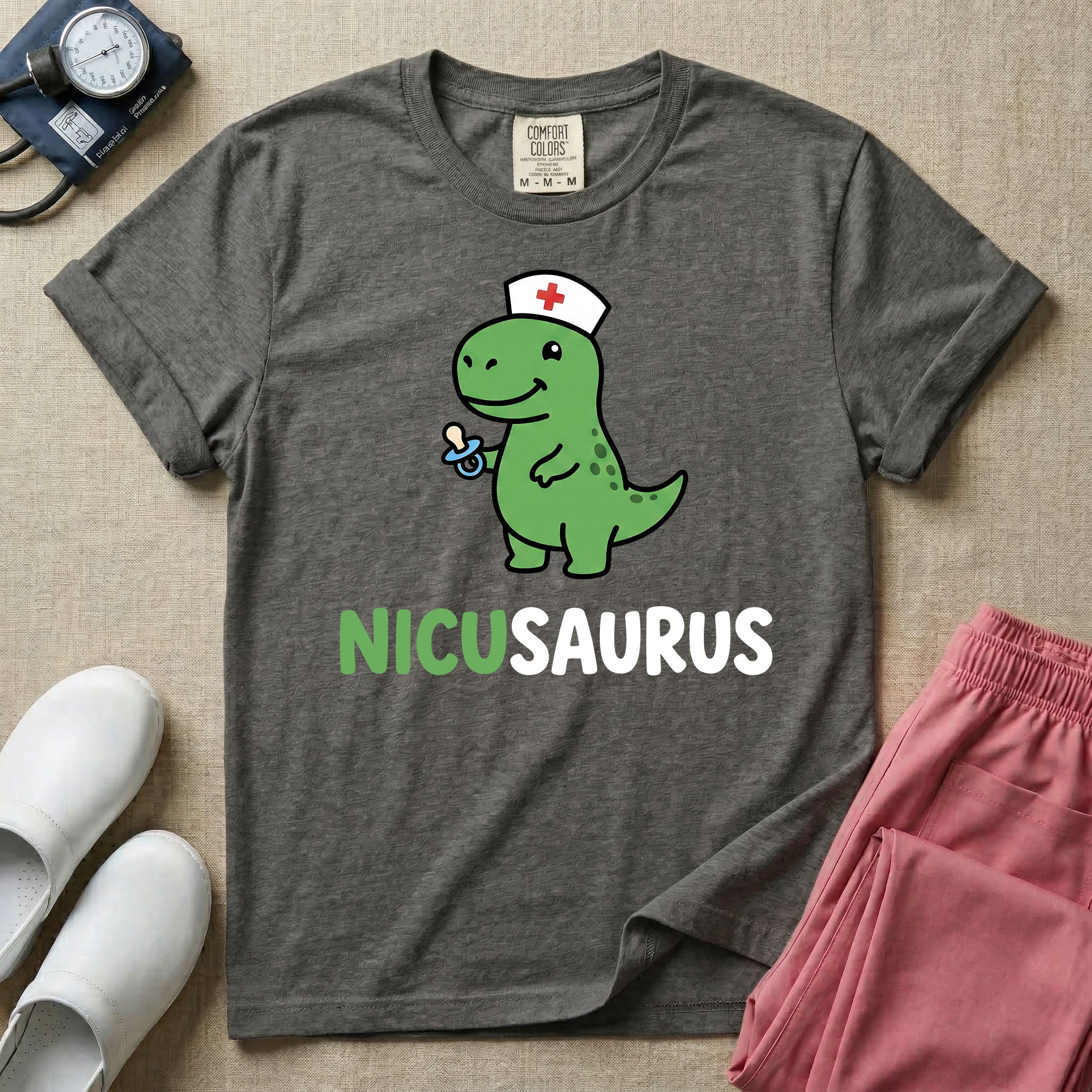 A cute t-shirt for NICU nurses featuring a cartoon green dinosaur wearing a nurse hat with a red cross and holding a blue pacifier above the funny text NICUSAURUS.