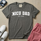 A minimalist personalized t-shirt for the NICU unit featuring bold white collegiate varsity lettering that reads NICU DAD arched over the text EST. 2026.