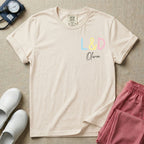 A personalized Labor and Delivery nurse t-shirt featuring a pastel front chest print with the letters L&D above a custom script name Olivia, and a large back print showcasing a cute cartoon stork flying with a baby bundle surrounded by small hearts and the humorous text Womb Service We Deliver.
