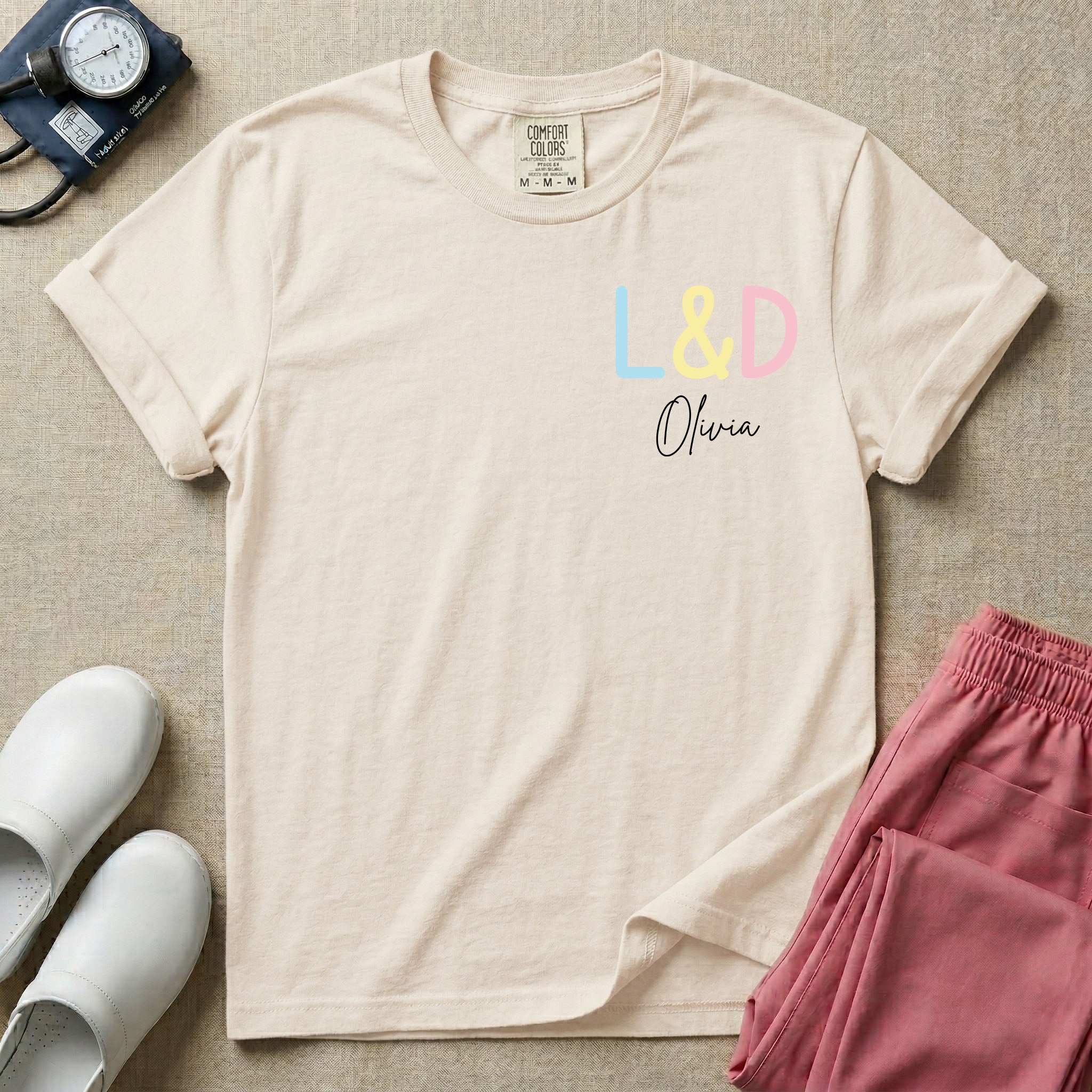 A personalized Labor and Delivery nurse t-shirt featuring a pastel front chest print with the letters L&D above a custom script name Olivia, and a large back print showcasing a cute cartoon stork flying with a baby bundle surrounded by small hearts and the humorous text Womb Service We Deliver.