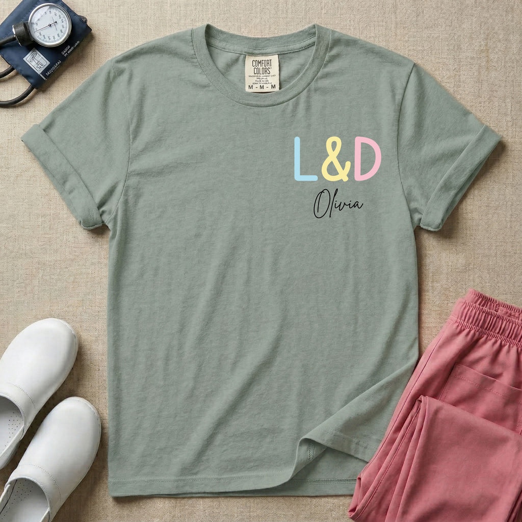 A personalized Labor and Delivery nurse t-shirt featuring a pastel front chest print with the letters L&D above a custom script name Olivia, and a large back print showcasing a cute cartoon stork flying with a baby bundle surrounded by small hearts and the humorous text Womb Service We Deliver.
