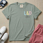 A personalized Labor and Delivery nurse t-shirt featuring a pastel front chest print with the letters L&D above a custom script name Olivia, and a large back print showcasing a cute cartoon stork flying with a baby bundle surrounded by small hearts and the humorous text Womb Service We Deliver.