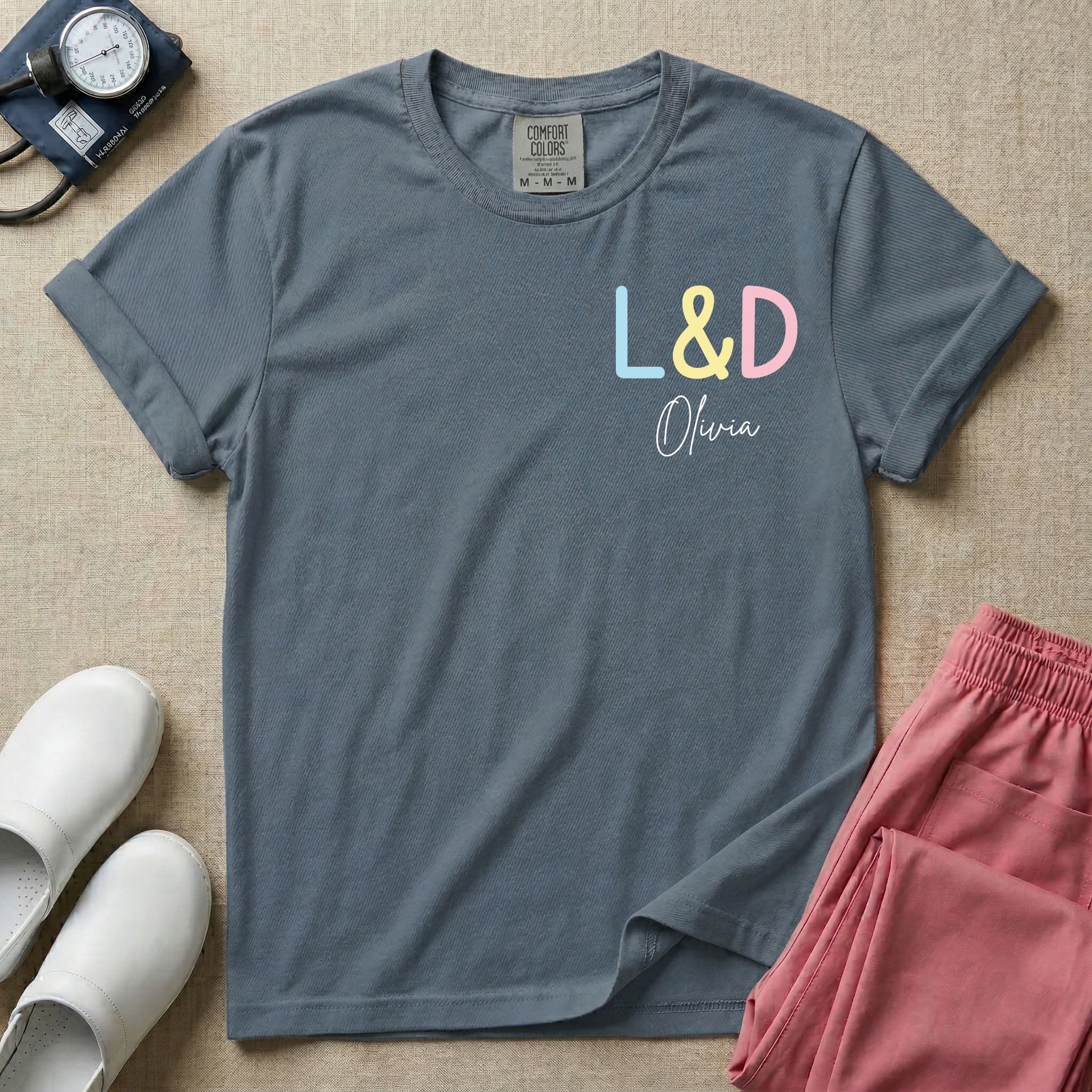A personalized Labor and Delivery nurse t-shirt featuring a pastel front chest print with the letters L&D above a custom script name Olivia, and a large back print showcasing a cute cartoon stork flying with a baby bundle surrounded by small hearts and the humorous text Womb Service We Deliver.