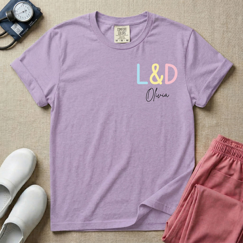 A personalized Labor and Delivery nurse t-shirt featuring a pastel front chest print with the letters L&D above a custom script name Olivia, and a large back print showcasing a cute cartoon stork flying with a baby bundle surrounded by small hearts and the humorous text Womb Service We Deliver.