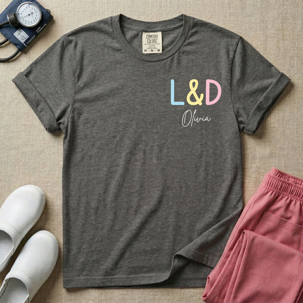 A personalized Labor and Delivery nurse t-shirt featuring a pastel front chest print with the letters L&D above a custom script name Olivia, and a large back print showcasing a cute cartoon stork flying with a baby bundle surrounded by small hearts and the humorous text Womb Service We Deliver.