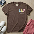 A personalized Labor and Delivery nurse t-shirt featuring a pastel front chest print with the letters L&D above a custom script name Olivia, and a large back print showcasing a cute cartoon stork flying with a baby bundle surrounded by small hearts and the humorous text Womb Service We Deliver.