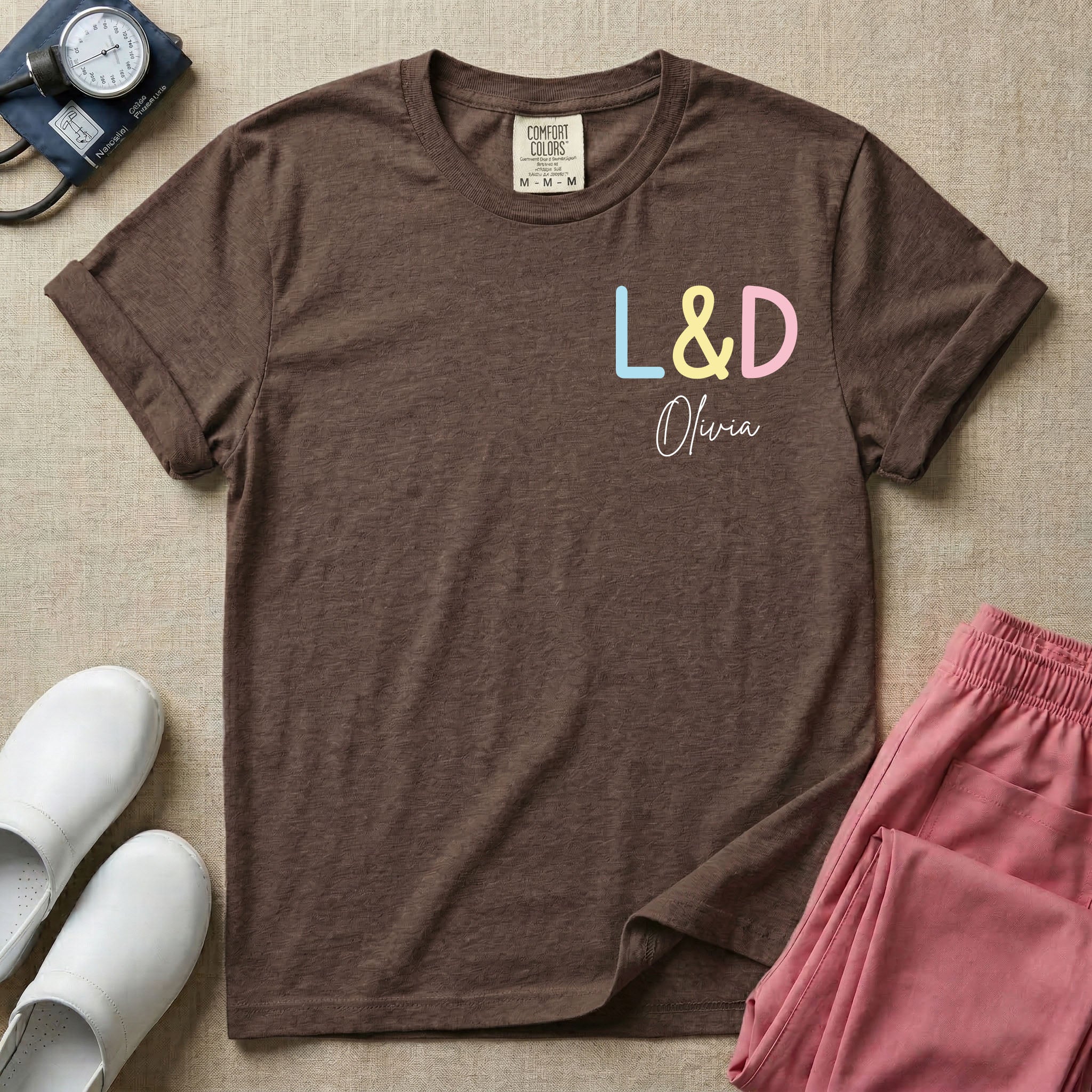 A personalized Labor and Delivery nurse t-shirt featuring a pastel front chest print with the letters L&D above a custom script name Olivia, and a large back print showcasing a cute cartoon stork flying with a baby bundle surrounded by small hearts and the humorous text Womb Service We Deliver.