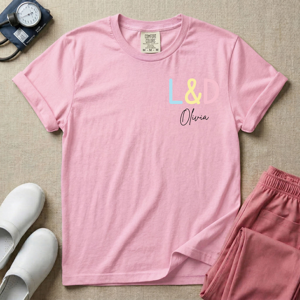 A personalized Labor and Delivery nurse t-shirt featuring a pastel front chest print with the letters L&D above a custom script name Olivia, and a large back print showcasing a cute cartoon stork flying with a baby bundle surrounded by small hearts and the humorous text Womb Service We Deliver.