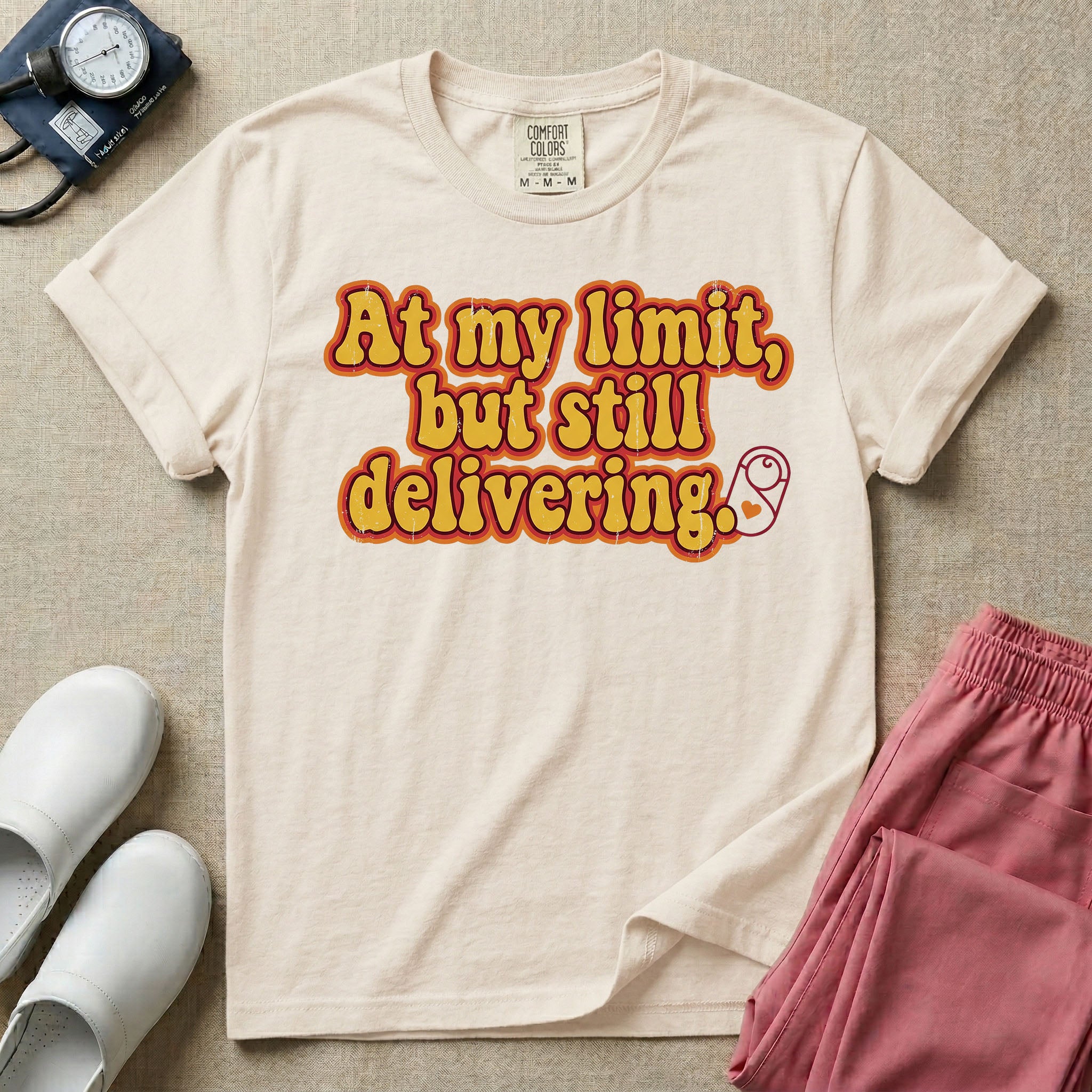 A retro-style Labor and Delivery nurse t-shirt featuring bold groovy typography that reads At my limit, but still delivering accompanied by a small minimalist graphic of a swaddled baby.