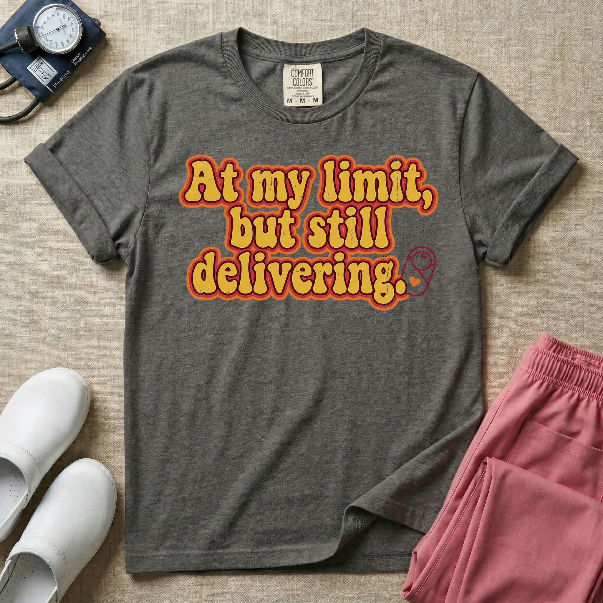 A retro-style Labor and Delivery nurse t-shirt featuring bold groovy typography that reads At my limit, but still delivering accompanied by a small minimalist graphic of a swaddled baby.