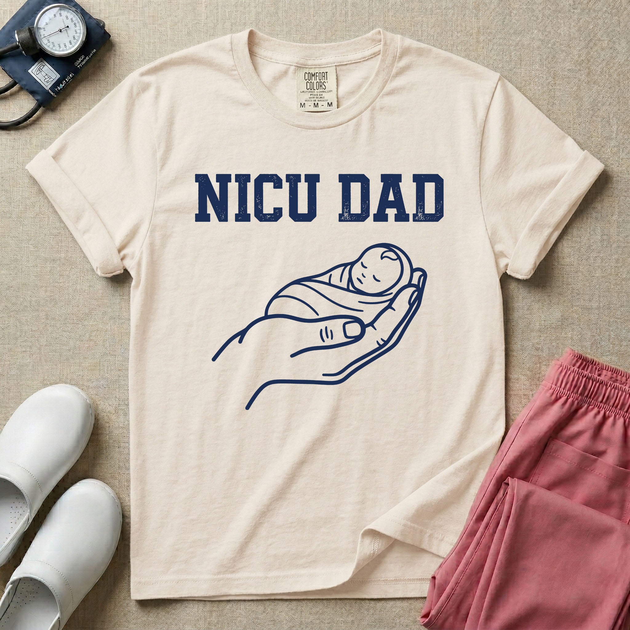 A minimalist NICU design featuring bold varsity text that reads NICU DAD above a gentle line art drawing of a large hand cradling a swaddled sleeping baby.