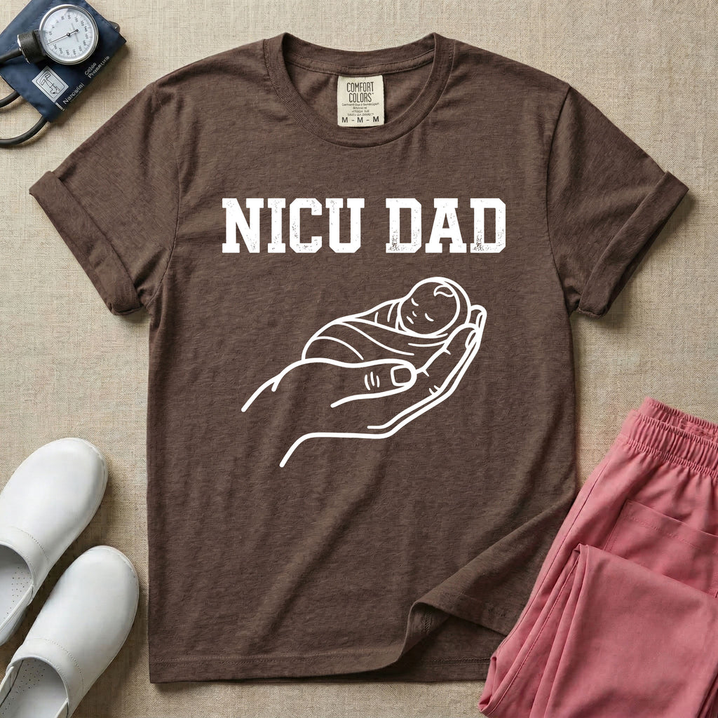 A minimalist NICU design featuring bold varsity text that reads NICU DAD above a gentle line art drawing of a large hand cradling a swaddled sleeping baby.