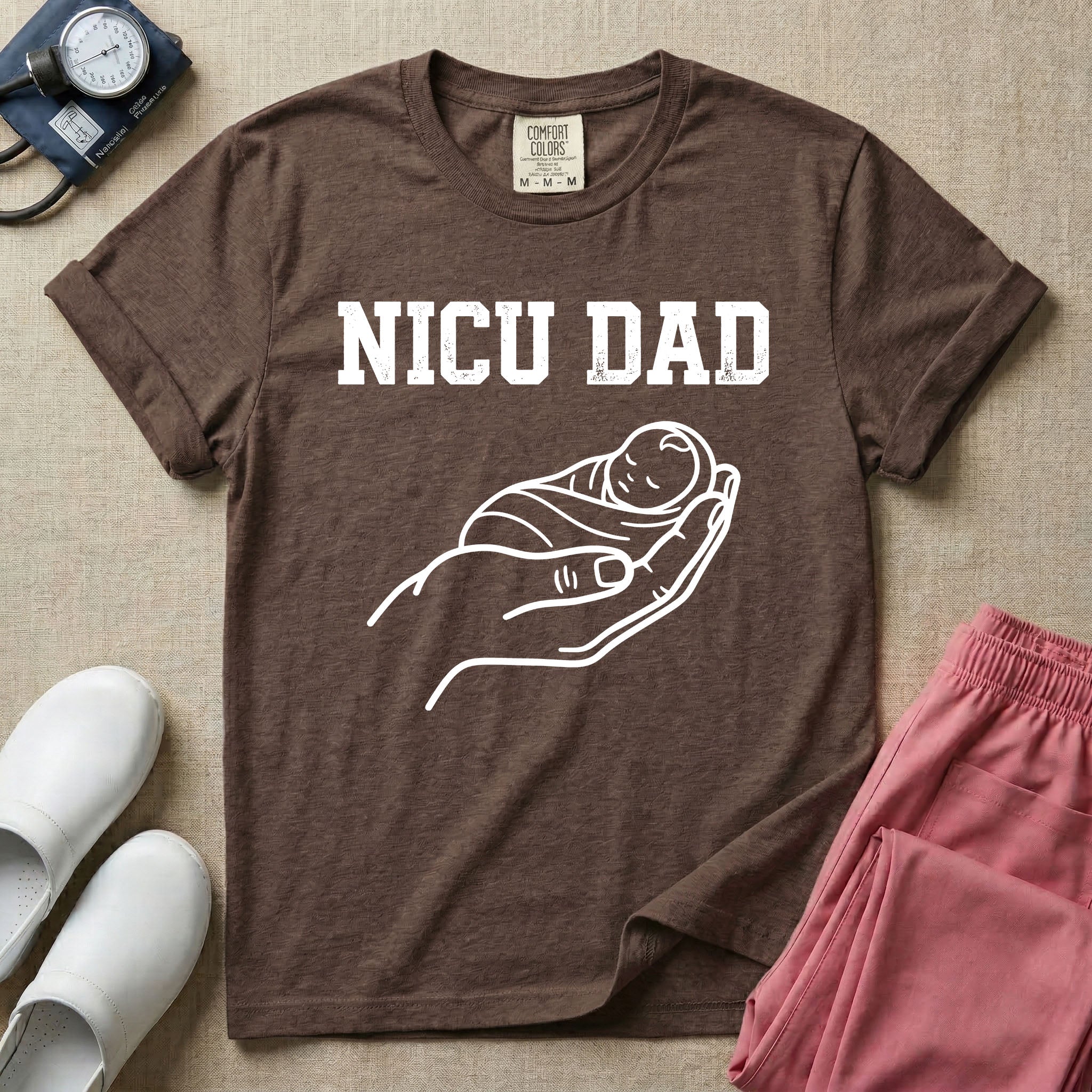 A minimalist NICU design featuring bold varsity text that reads NICU DAD above a gentle line art drawing of a large hand cradling a swaddled sleeping baby.