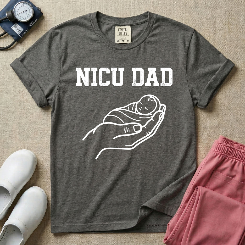A minimalist NICU design featuring bold varsity text that reads NICU DAD above a gentle line art drawing of a large hand cradling a swaddled sleeping baby.