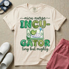 Cute and funny NICU nurse design featuring the text Incu-Gator Tiny But Mighty with an adorable baby alligator swaddled in a hospital receiving blanket resting inside an incubator isolette.