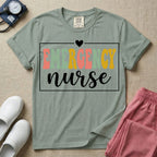 A retro-style t-shirt for emergency room nurses featuring the word "EMERGENCY" in colorful, vintage lettering above the word "nurse" in a delicate white script font, framed by a thin white rectangular border with a small heart at the top center.