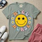A t-shirt featuring a large yellow smiley face with star eyes surrounded by smiley face daisies and the text N.I.C.U. NURSE in pastel retro bubble lettering.
