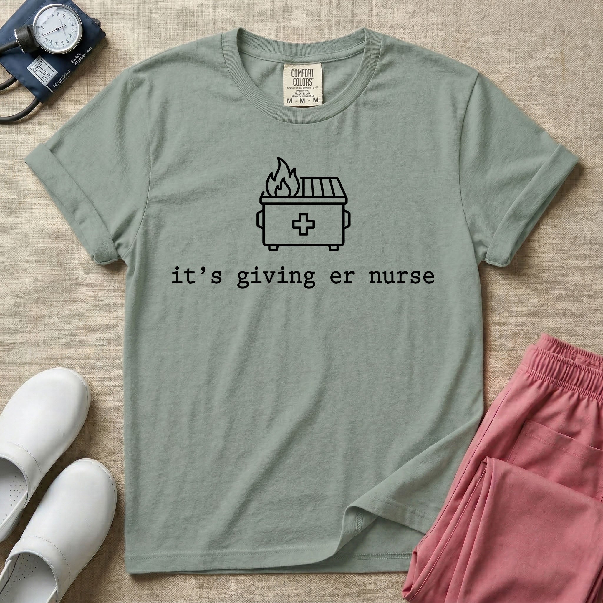 A funny minimalist ER nursing t-shirt featuring a simple white line drawing of a dumpster on fire with a medical cross on it, sitting above the lower-case typewriter text "it's giving er nurse".