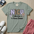 A funny t-shirt for NICU nurses featuring the text "NICU I Work for a 4-Pound Dictator" designed with purple plaid and leopard print letters, accompanied by graphics of a baby bottle with a bow and a rattle that says "WARNING: TINY BOSS".
