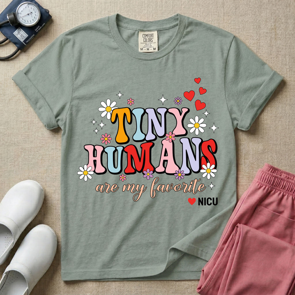 A t-shirt featuring the text Tiny Humans are my favorite in colorful retro bubble letters decorated with daisies, sparkles, and red hearts with a small NICU heart logo at the bottom.