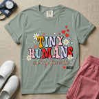 A t-shirt featuring the text Tiny Humans are my favorite in colorful retro bubble letters decorated with daisies, sparkles, and red hearts with a small NICU heart logo at the bottom.