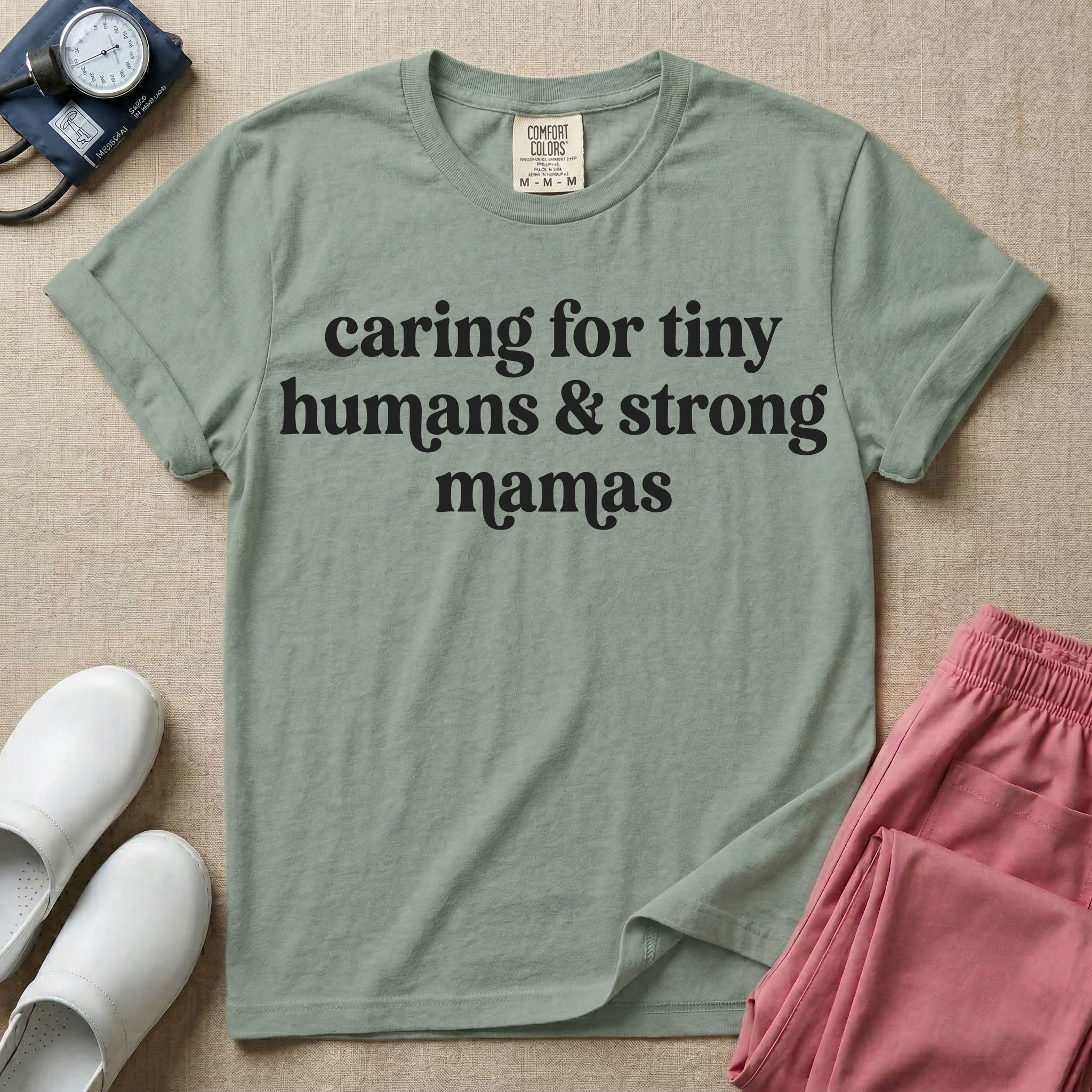 A minimalist nursing t-shirt featuring the text "caring for tiny humans & strong mamas" written in a retro white serif font.