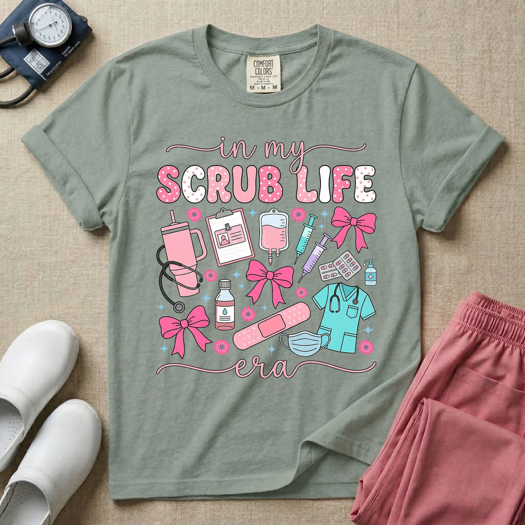 A cute general nursing t-shirt featuring the retro text "in my SCRUB LIFE era" surrounded by coquette medical graphics including a pink tumbler cup, stethoscope, IV bag with pink bows, clipboard, and blue scrubs.