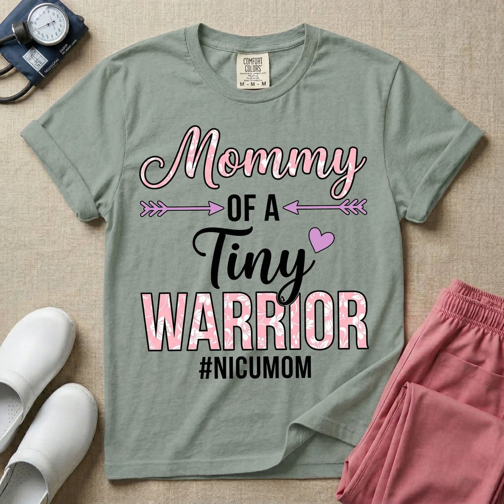 A cute NICU mom t-shirt featuring the stacked text "Mommy OF A Tiny WARRIOR #NICUMOM" in pink and white, accented with decorative arrows, a small pink heart, and a pink plaid pattern filling the word warrior.