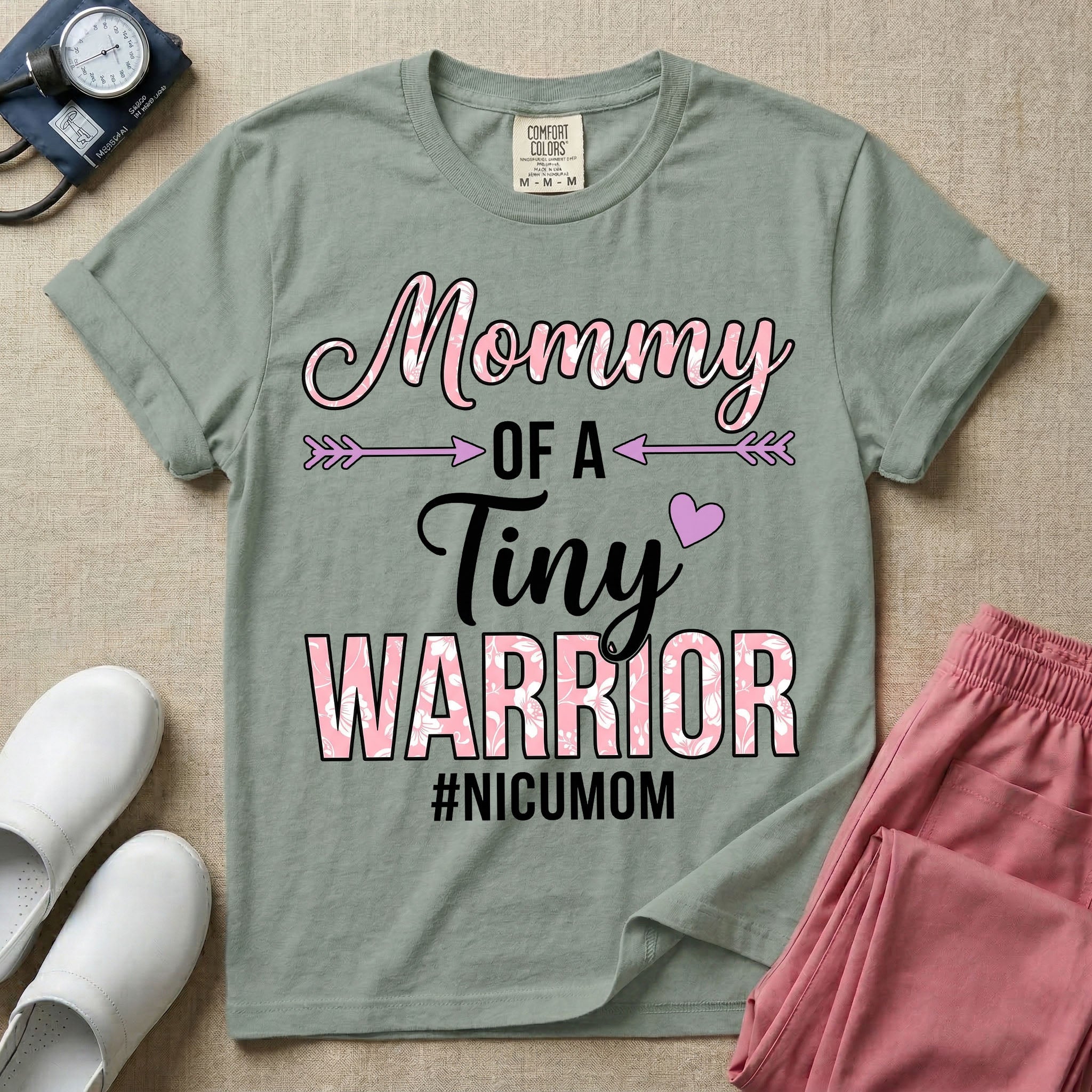 A cute NICU mom t-shirt featuring the stacked text "Mommy OF A Tiny WARRIOR #NICUMOM" in pink and white, accented with decorative arrows, a small pink heart, and a pink plaid pattern filling the word warrior.