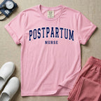 A t-shirt featuring the word POSTPARTUM arched in large collegiate block letters above the word NURSE in a smaller, matching font.