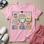 A retro-style t-shirt for emergency room nurses featuring the text "ER CREW" below a vintage cartoon illustration of a medicine bottle, a grumpy clipboard holding an AMA form, and a sandwich wearing a nurse cap walking together.