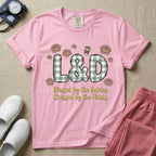 A descriptive graphic tee for labor and delivery nurses featuring plaid block letters spelling L&D surrounded by floral elements and a coffee cup, with the text "Blessed by the Babies, Stressed by the Fluids."