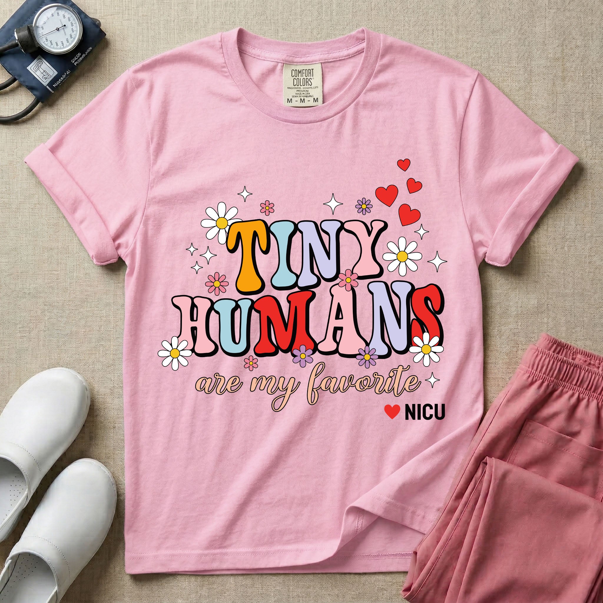A t-shirt featuring the text Tiny Humans are my favorite in colorful retro bubble letters decorated with daisies, sparkles, and red hearts with a small NICU heart logo at the bottom.