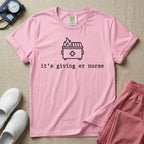 A funny minimalist ER nursing t-shirt featuring a simple white line drawing of a dumpster on fire with a medical cross on it, sitting above the lower-case typewriter text "it's giving er nurse".