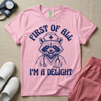 A humorous graphic of a raccoon dressed in a nursing cap and scrub jacket with a stethoscope, with the text "FIRST OF ALL I'M A DELIGHT", in blossom color.