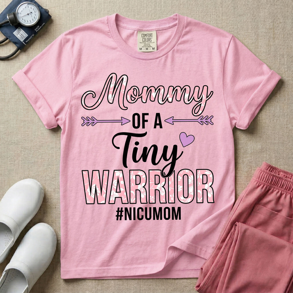 A cute NICU mom t-shirt featuring the stacked text "Mommy OF A Tiny WARRIOR #NICUMOM" in pink and white, accented with decorative arrows, a small pink heart, and a pink plaid pattern filling the word warrior.