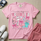A cute general nursing t-shirt featuring the retro text "in my SCRUB LIFE era" surrounded by coquette medical graphics including a pink tumbler cup, stethoscope, IV bag with pink bows, clipboard, and blue scrubs.