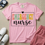 A retro-style t-shirt for emergency room nurses featuring the word "EMERGENCY" in colorful, vintage lettering above the word "nurse" in a delicate white script font, framed by a thin white rectangular border with a small heart at the top center.