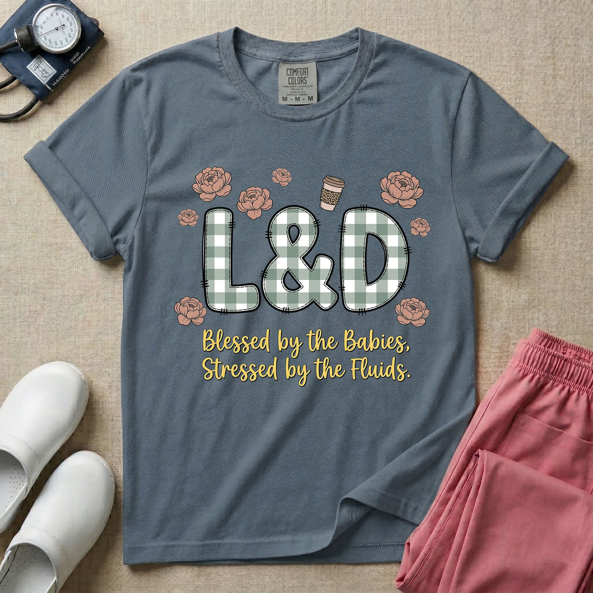 A descriptive graphic tee for labor and delivery nurses featuring plaid block letters spelling L&D surrounded by floral elements and a coffee cup, with the text "Blessed by the Babies, Stressed by the Fluids."