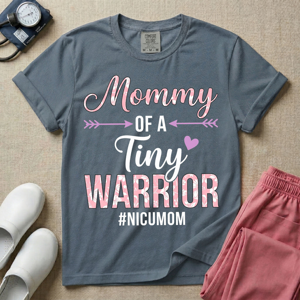 A cute NICU mom t-shirt featuring the stacked text "Mommy OF A Tiny WARRIOR #NICUMOM" in pink and white, accented with decorative arrows, a small pink heart, and a pink plaid pattern filling the word warrior.