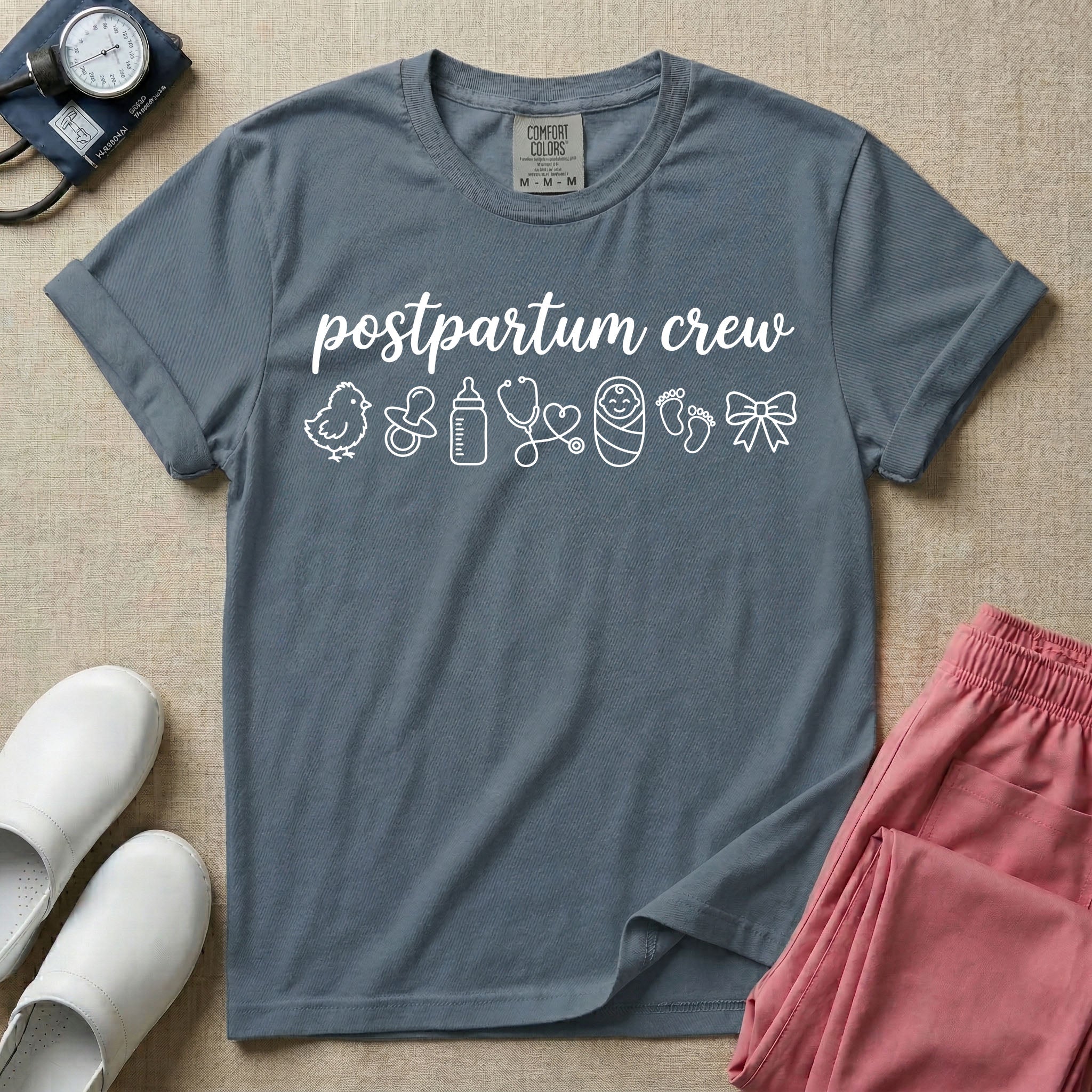 A t-shirt featuring the lowercase text postpartum crew in a cursive script font above a horizontal row of minimalist line-art icons including a baby chick, a pacifier, a baby bottle, a heart-shaped stethoscope, a swaddled baby, baby footprints, and a ribbon bow.