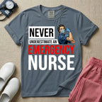 A retro ER nursing t-shirt featuring the bold text "NEVER UNDERESTIMATE AN EMERGENCY NURSE" alongside a vintage graphic of a masked nurse flexing her arm in a classic Rosie the Riveter pose