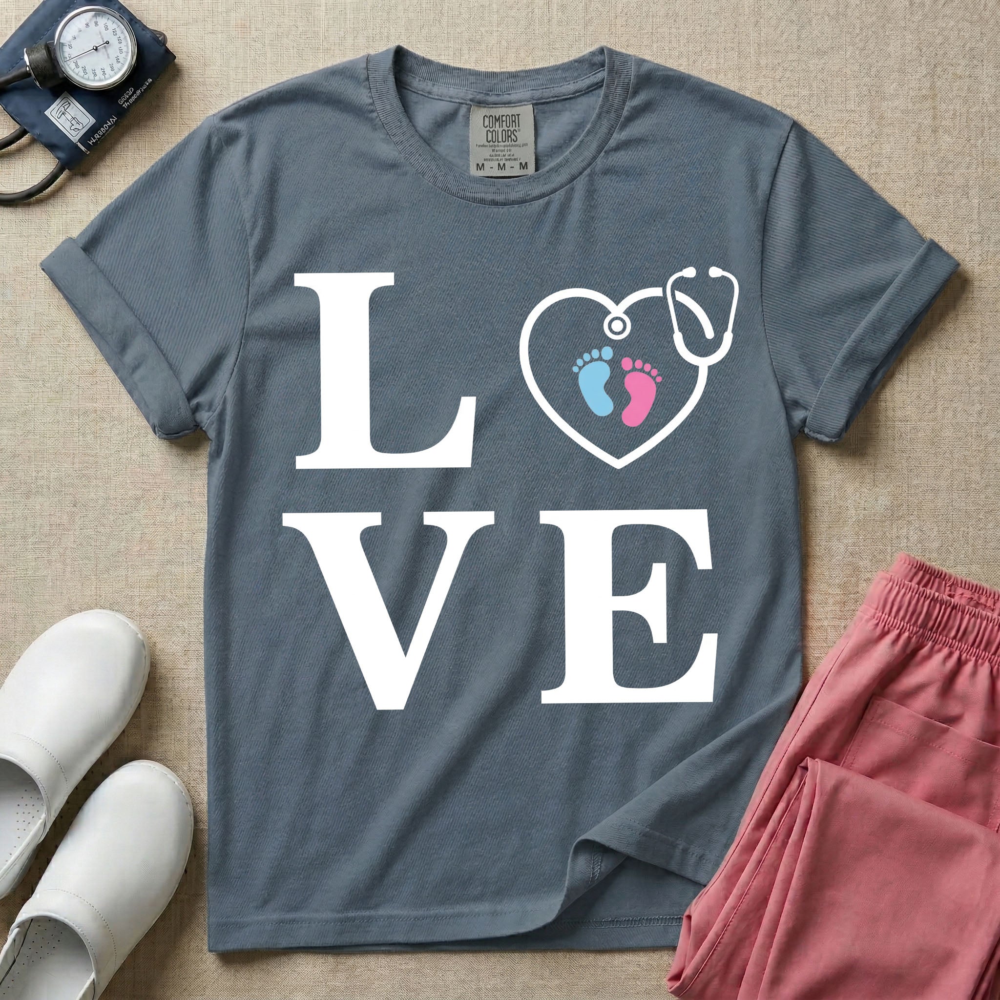 A minimalist nursing t-shirt featuring the word LOVE stacked in large white serif letters, with the letter O replaced by a white stethoscope shaped like a heart containing two small baby footprints, one pink and one blue.
