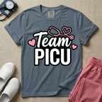 A descriptive graphic tee for pediatric intensive care nurses featuring the text "Team PICU" in bold white lettering, accented with pink hearts and a pink stethoscope design.