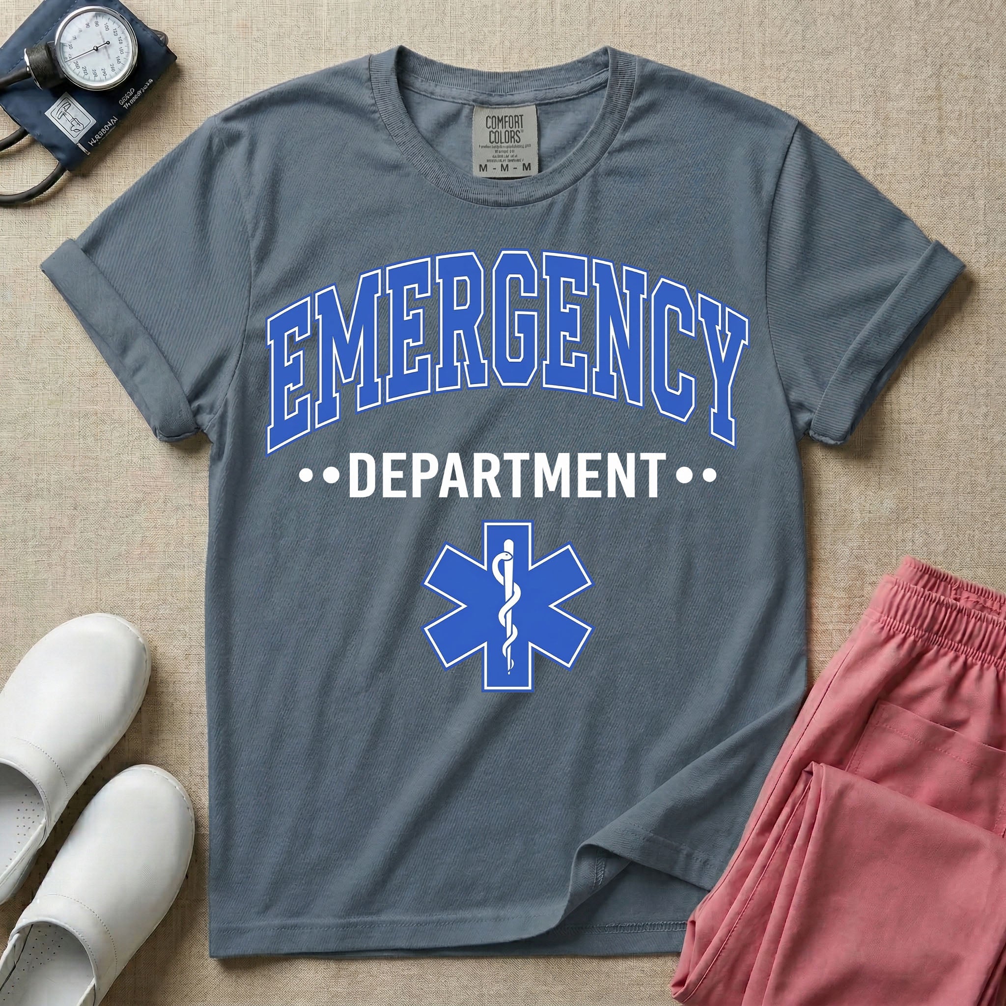 A classic varsity-style t-shirt for emergency room nurses featuring the word "EMERGENCY" in a large, arched blue collegiate font above the word "DEPARTMENT", centered over a blue Star of Life medical symbol.