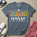 A retro-style t-shirt for emergency room nurses featuring the word "EMERGENCY" in colorful, vintage lettering above the word "nurse" in a delicate white script font, framed by a thin white rectangular border with a small heart at the top center.