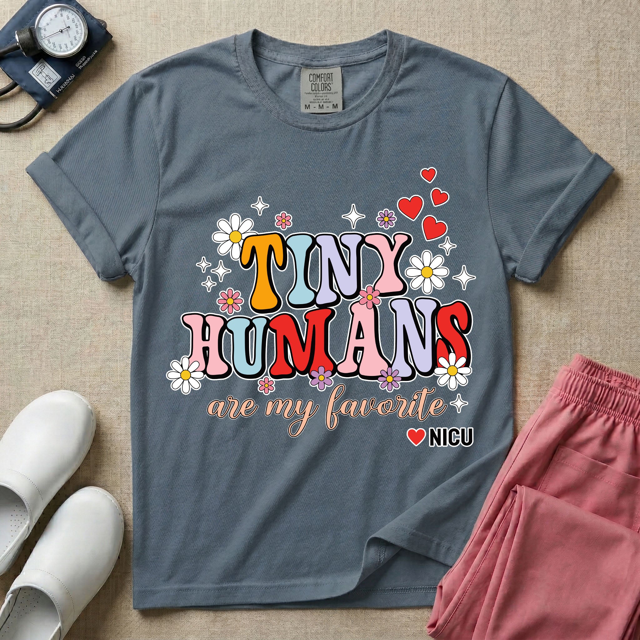 A t-shirt featuring the text Tiny Humans are my favorite in colorful retro bubble letters decorated with daisies, sparkles, and red hearts with a small NICU heart logo at the bottom.
