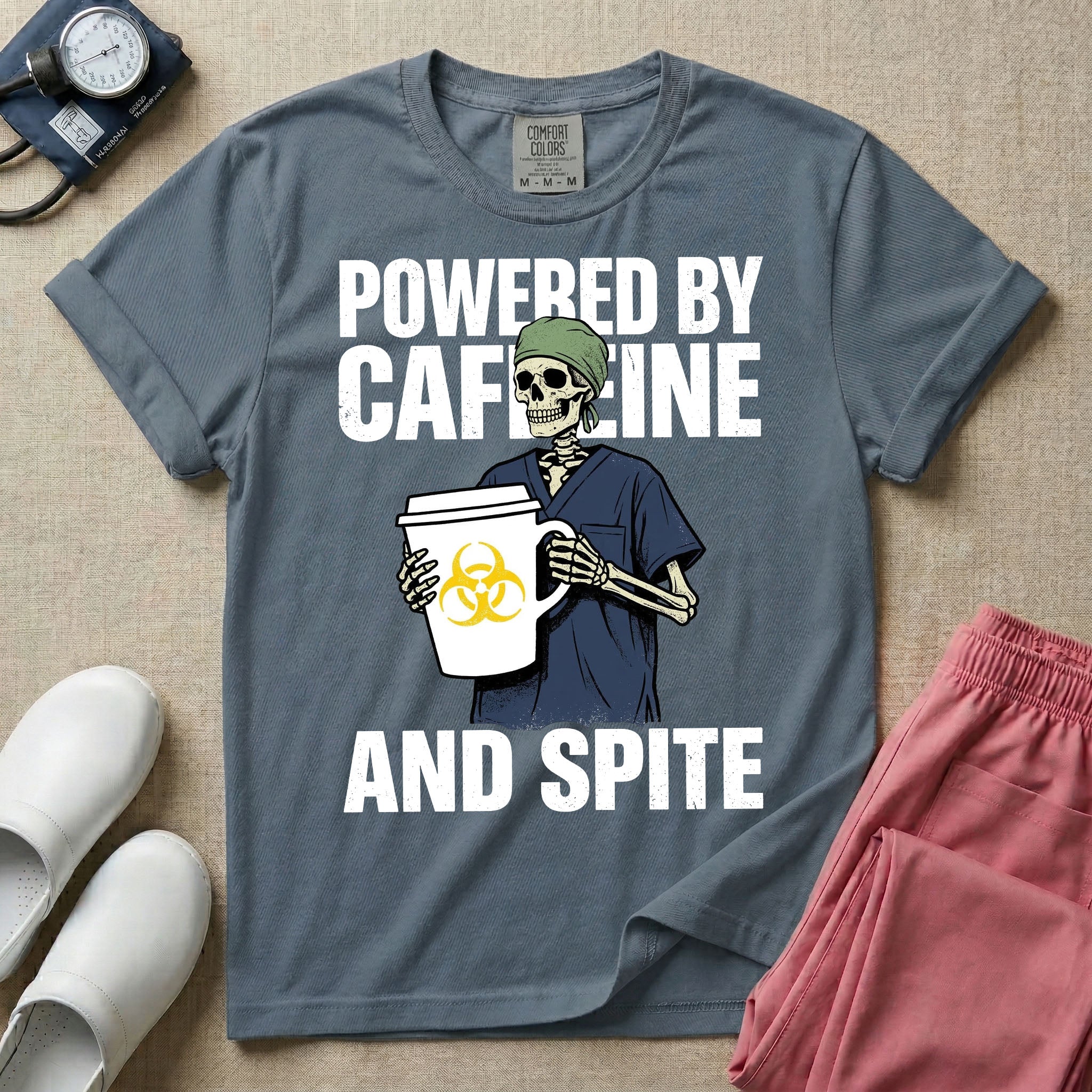A humorous graphic for general nursing featuring an illustration of a skeleton wearing a green scrub cap and dark blue scrubs, holding a large white coffee cup with a yellow biohazard symbol, with the stacked text 'POWERED BY CAFFEINE' above it and 'AND SPITE' below it.