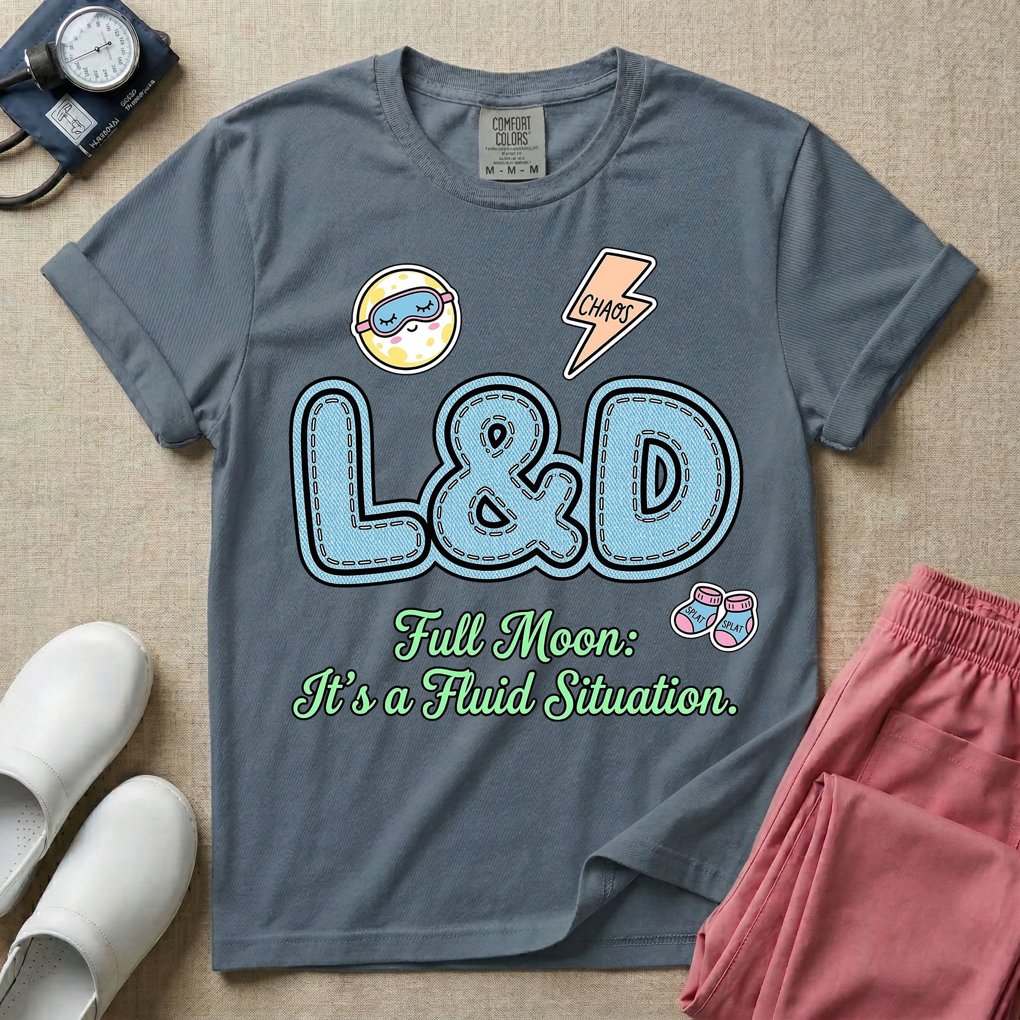 A humorous Labor and Delivery nurse graphic featuring large, light blue varsity-style letters spelling L&D with a pink dashed outline, surrounded by small patch-like icons of a baby and a lightning bolt, sitting above cursive text that reads Full Moon: It's a Fluid Situation.