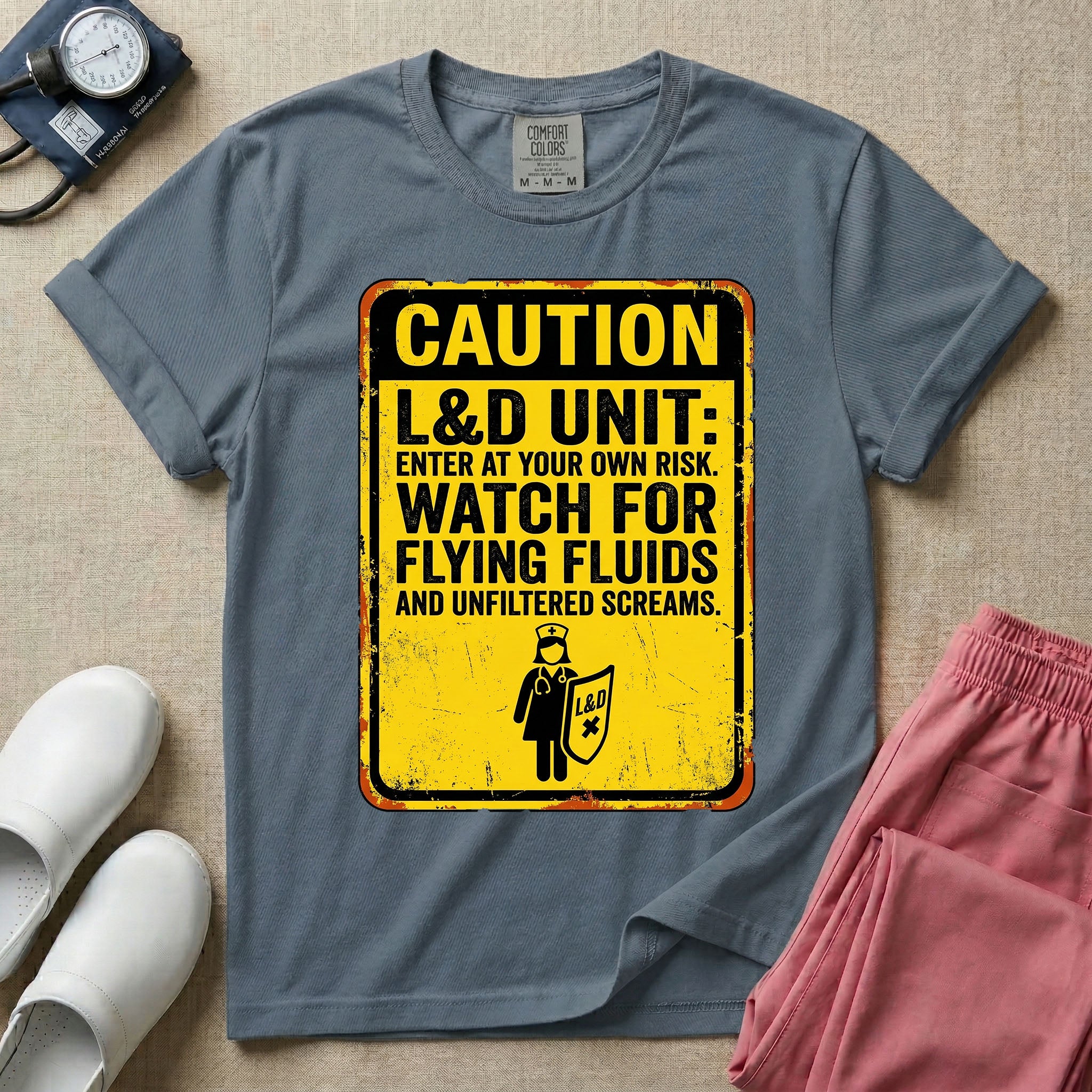 A humorous graphic tee for labor and delivery nurses featuring a distressed caution sign design with the text "CAUTION L&D UNIT: ENTER AT YOUR OWN RISK. WATCH FOR FLYING FLUIDS AND UNFILTERED SCREAMS." above a stick figure nurse holding a shield.